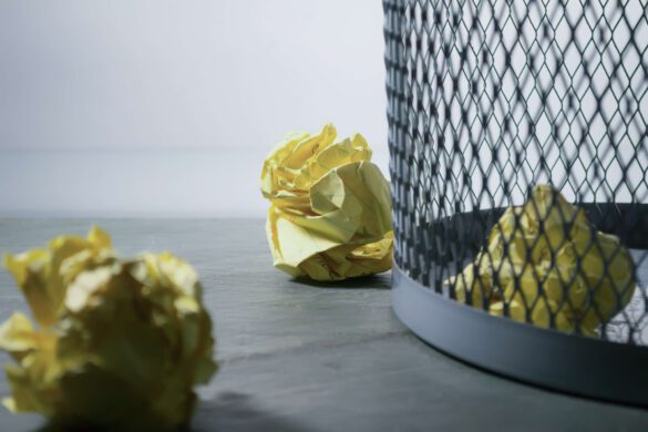 3 zgięte kulki z papieru, z których jedna leży w koszu na śmieci, a dwie leżą tuż obok kosza na śmieci