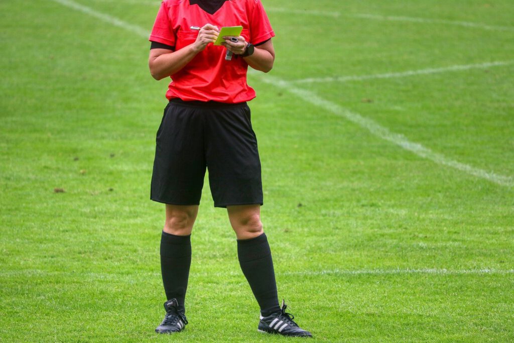 Sędzia ubrany w czerwoną koszulkę, czarne spodenki i getry - stoi na murawie piłkarskiej zapisuje coś na żółtej kartce.