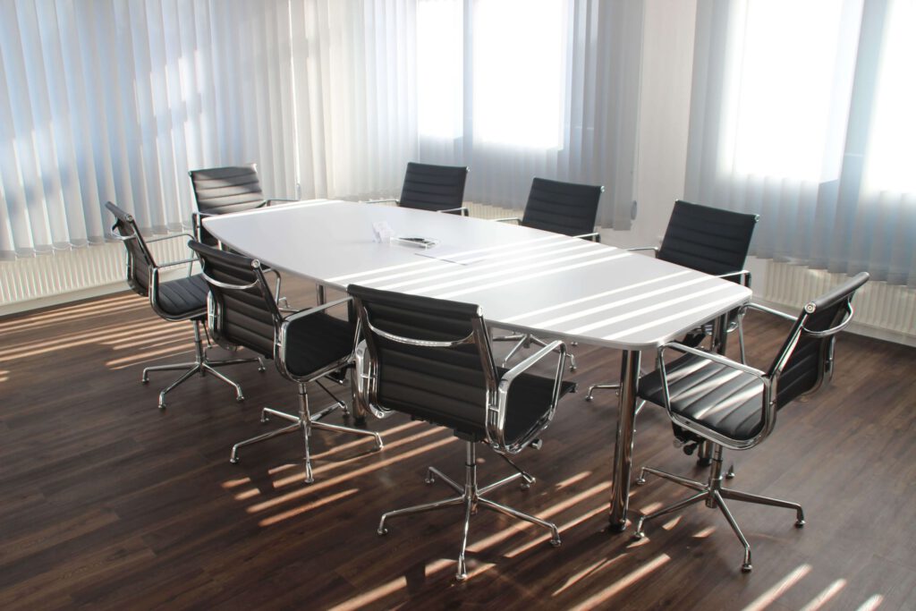 Sala konferencyjna, na środku której stoi owalny biały stół z ośmioma krzesłami biurowymi. W tle widać ściany i okna zakryte roletami.