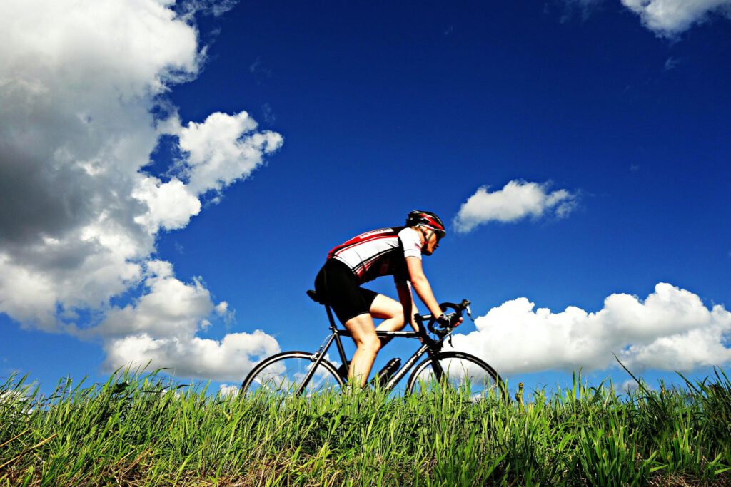 Mężczyzna w stroju kolarskim i kasku jadący na rowerze przy trawie. W tle błękitne niebo z kilkoma chmurami.